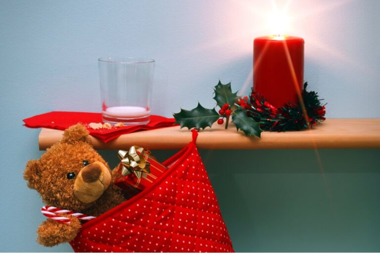 Christmas stocking holding a teddy bear hangning from shelf with lit candle and empty glass of milk